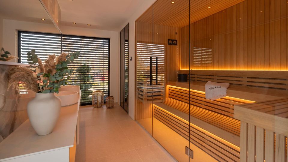 Moderner Spa- bzw. Saunaraum mit großflächiger Glasfront zur holzverkleideten Sauna, innen mit beleuchteten Holz-Liegen und warmen LED-Lichtleisten. Helle Fliesen, große Fenster mit Lamellenjalousien und eine Ablage mit Vasen und Trockenpflanzen prägen den Raum.