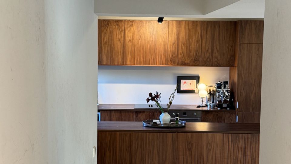 View down a narrow white-walled hallway into a modern kitchen with walnut wood cabinets and a matching island, lit by a small overhead spotlight. A high rectangular window near the ceiling brings in daylight above the kitchen area.