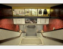 Treppe in einer U-Bahn-Station mit rot-gemusterten Wänden, Geländern aus Holz und Betonstufen. Oben befindet sich ein Schaufenster mit Ladenansicht, und an der Wand hängt eine mit Graffiti versehene Werbung.