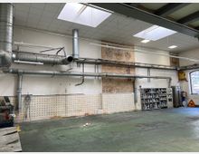 Industriehalle mit hohen Decken, sichtbaren Lüftungsrohren und Oberlichtern. Wände mit teilweise unverputztem Backstein, Regalen voller Behälter und großen Fenstern, die Tageslicht hereinlassen.