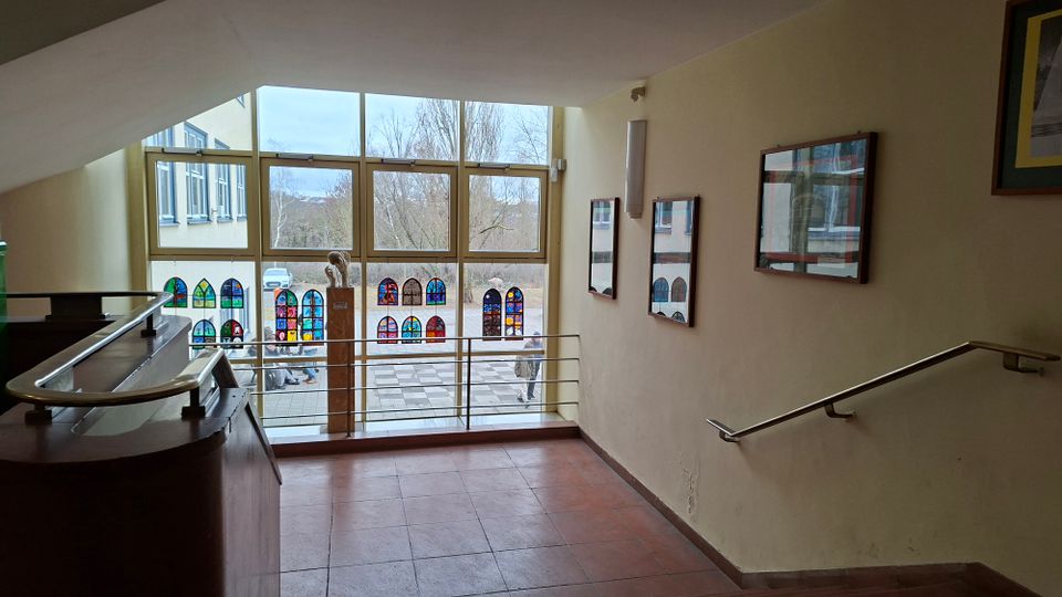 Helles Treppenhaus oder Flur mit Fliesenboden und Handläufen, das zu einer großen Fensterfront führt. Vor den Fenstern sind bunte, bleiglasartige Dekore angebracht; an den Wänden hängen gerahmte Bilder, draußen sind Gebäude und Bäume sichtbar.