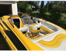 Gelbes und weißes Motorboot mit offenem Cockpit und gepolsterten Sitzen, geparkt vor einer Garage mit einer Säule. Im Hintergrund ist ein Garten mit grüner Rasenfläche und Bäumen sowie ein modernes Haus zu sehen.