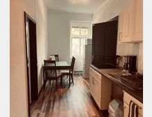 Schmale Wohnküche mit Holzoptikboden, Esstisch und vier Stühlen vor einem großen Fenster. Rechts eine weiße Küchenzeile mit Spüle und Arbeitsplatte sowie ein hoher Kühlschrank neben einer dunklen Tür.