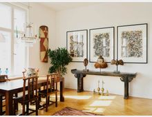 Esszimmer mit Holzmöbeln, darunter ein großer rechteckiger Tisch und Stühle. An der Wand hängen drei abstrakte Kunstwerke, daneben steht ein Konsolentisch mit dekorativen Masken. Ein Kronleuchter hängt von der Decke, und auf dem Holzboden liegt ein Teppich. Ein großes Fenster lässt Tageslicht herein und eine Pflanze ergänzt den Raum.