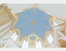 Blick auf eine blaue, gewölbte Kirchendecke mit goldenen Sternen und Rippenbögen. Darunter befinden sich hohe bunte Glasfenster und ein großer, runder Kronleuchter in der Mitte.