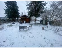 Schneebedeckter Garten mit einer kleinen Holzhütte im Hintergrund, umgeben von Bäumen, Sträuchern und einem Zaun. Im Vordergrund stehen eine Holzbank, ein Tisch und verstreute Gartenmöbel.