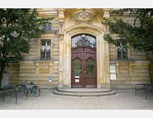 Eingang eines historischen Gebäudes mit großen, verzierten Holztüren und einem geschwungenen Steinportal, flankiert von Fenstern mit schmiedeeisernen Gittern. Vor dem Gebäude steht ein Fahrradständer mit zwei Fahrrädern, und ein Schild für barrierefreien Zugang ist sichtbar.