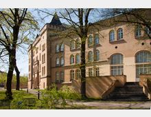 Historisches Backsteingebäude mit Spitzdach und Rundbogenfenstern, umgeben von Bäumen in einer parkähnlichen Landschaft; eine breite Treppe führt zum Eingangsbereich.
