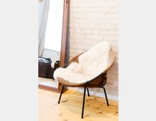 Ein Wohnzimmer mit hellem Holzboden, einer gemauerten Ziegelwand, einem braunen Ledersessel mit heller Fellauflage und einem großen Holzrahmenspiegel, der an die Wand gelehnt ist. Im Hintergrund ist ein wenig Licht durch ein Fenster zu sehen.