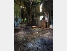 Innenraum eines verlassenen Gebäudes mit stark beschädigten Wänden und Decke. Der Raum ist dunkel, mit kaputten Fenstern, freiliegenden Heizkörpern und einem Boden voller Schutt und Moos.