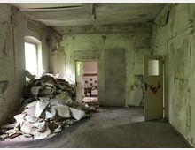 Ein heruntergekommenes, leerstehendes Zimmer mit abblätternden Wänden, einem Haufen von Schutt und Tapetenresten auf dem Boden sowie einer offenen Tür, die in einen Raum mit weiß-gelben Fliesen führt. Die Atmosphäre wirkt vernachlässigt, mit begrenztem Tageslicht, das durch ein Fenster einfällt.