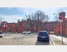 Backsteinbau mit neugotischen Elementen und einem Torbogen, darüber der Schriftzug „Technische Hochschule Brandenburg“. Im Vordergrund eine gepflasterte Straße mit parkenden Autos und einem Verkehrsschild, im Hintergrund Bäume und der Hauptgebäudekomplex.