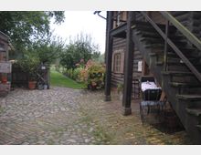 Ein rustikaler Hof mit einem Holzgebäude, einer Außentreppe und einer gepflasterten Fläche. Im Hintergrund sind ein Garten mit Bäumen und Sträuchern sowie eine schmale Wegführung erkennbar.