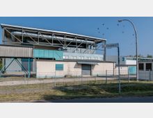 Industriegebäude mit flachem Dach und sichtbaren Rohren auf einer oberen Ebene, umgeben von einem Zaun. Im Vordergrund verläuft eine Straße, daneben ein Straßenschild mit der Aufschrift „Zum Umspannwerk 4“.