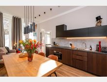 Offene, moderne Wohnküche mit langem Holztisch und hängenden schwarzen Pendelleuchten, daneben eine Küchenzeile mit dunklen Holzfronten, schwarzen Oberschränken und grauer Rückwand mit Unterbauleiste. Große Fenster mit grauen Vorhängen und Holzfußboden sorgen für viel Tageslicht im Raum.