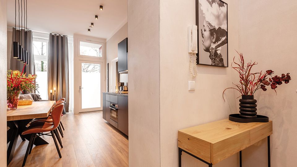 Heller, moderner Wohn- und Essbereich mit langem Holztisch und Lederstühlen, schwarzer Küchenzeile sowie hohen Fenstern und Glastür mit grauen Vorhängen. Im Vordergrund steht eine schmale Holz-Konsole mit Vase und Wandbild an einer weißen Trennwand; Holzfußboden und Deckenspots prägen den Raum.