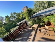 Holzterrasse mit Geländer, umgeben von grünen Bäumen und Sträuchern, im Hintergrund ein Flachdachgebäude mit Schornstein. Auf der Terrasse stehen einfache Holzmöbel, darunter ein Tisch und zwei Stühle, teils im Schatten.