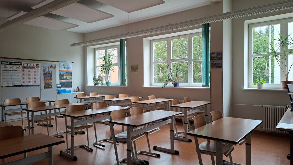 Heller Klassenraum mit mehreren Reihen aus Holztischen und Stühlen sowie großen Fenstern an der Längsseite mit grünen Vorhängen und Pflanzen auf den Fensterbänken. An den Wänden hängen Unterrichtsplakate, und an der Decke sind rechteckige Akustikpaneele und Leuchtleisten montiert.