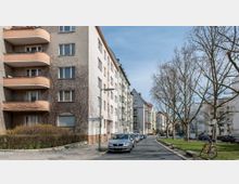 Straßenszene in einem Wohngebiet mit mehrstöckigen Apartmentgebäuden, Balkonen und geparkten Autos am Straßenrand. Im Vordergrund Gehwege, karge Bäume und ein Fahrrad an einem Poller, bei klarem Himmel.