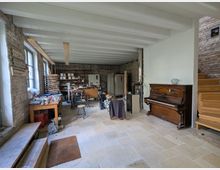 Ein Werkstattraum mit offener, rustikaler Gestaltung, gekennzeichnet durch unverputzte Ziegelwände, sichtbare Holzträger an der Decke und große Fenster. Im Raum befinden sich Arbeitsflächen mit Werkzeugen, Regale mit Materialien, ein Holzofen, ein antikes Klavier und eine Holztreppe, die nach oben führt.