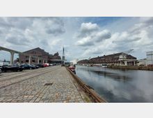 Pflasterstraße entlang eines Industriehafens mit roten Backsteingebäuden auf beiden Seiten des Kanals. Links sind parkende Autos und ein Schienensystem, rechts Kräne, Boote und eine spiegelnde Wasserfläche unter bewölktem Himmel.