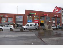 Außenansicht eines Einkaufszentrums an der Hermannstraße mit einer roten Backsteinfassade und Geschäften wie Rewe, Netto, dm-Drogeriemarkt und NKD. Vor dem Gebäude befinden sich parkende Autos, ein Lieferwagen mit der Aufschrift „MILES“ sowie eine U-Bahn-Station mit dem Schild „U Hermannstraße“.