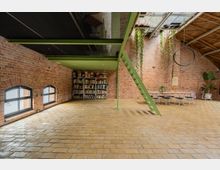 Industrie-inspirierter Innenraum mit Backsteinwänden, einem hellen Ziegelboden und einer grünen Metallstruktur, die eine Treppe und eine Zwischendecke umfasst. Links stehen Bücherregale, während rechts ein Esstisch mit Stühlen, hängenden Pflanzen und ein großer schwarzer Ring als Leuchte sichtbar sind.