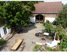 Ein gepflasterter Innenhof mit Sitzmöglichkeiten, einer Feuerstelle und viel Grün. Links steht ein großes Gebäude mit weißen Wänden und Ziegeln, rechts eine mit Rosen bewachsene Ecke, in der Mitte ein großer Baum.