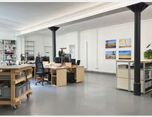 Heller, offener Büroraum mit Betonboden, weißen Wänden und schwarzen Stützsäulen, beleuchtet durch lange Deckenleuchten. Mehrere Holzschreibtische mit Computerarbeitsplätzen, Regale mit Büromaterial sowie an der Wand befestigte Fotodrucke und ein Metallregal prägen den Raum.