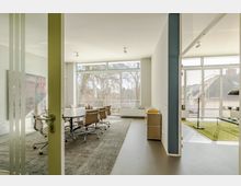 Ein moderner Konferenzraum mit einem großen rechteckigen Tisch und drehbaren Bürostühlen auf einem grauen Teppich. Der Raum hat große Fenster mit Jalousien, die Tageslicht hereinlassen, und im Hintergrund ist ein weiterer Bereich mit grüner Teppichfläche sichtbar.