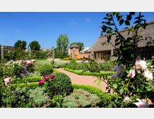Gartenanlage mit Rosenbeeten, gepflegten Hecken und geschwungenen Gehwegen, im Hintergrund ein Fachwerkgebäude mit Reetdach und ein Backsteinturm. Umgeben von Bäumen und einigen modernen Gebäuden unter blauem Himmel.