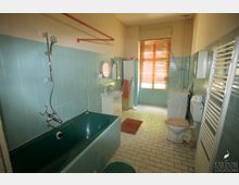 Badezimmer mit mintgrünen Wandfliesen, türkisfarbener Badewanne und weißem Fliesenboden. Das Zimmer verfügt über ein Waschbecken mit Spiegel, eine Toilette, eine Waschmaschine und ein großes Fenster mit Jalousien.