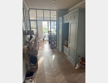 Ein schmaler Flur mit hellem Fischgrätparkettboden, rechts eingebauten graugrünen Schränken mit Sitzbank und links einer schmalen Holzkonsole mit Dekoration. Am Ende des Flurs befindet sich eine Glaswand mit schwarzem Metallrahmen, durch die ein helles Wohnzimmer mit großen Fenstern sichtbar ist.