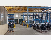 Innenansicht einer Werkshalle mit Industriemaschinen, blauen Stahlträgern und gelben Krananlagen an der Decke. Im Vordergrund sind Radsätze auf Paletten gestapelt, während Regale mit Metallteilen und Arbeitsstationen im Hintergrund sichtbar sind.