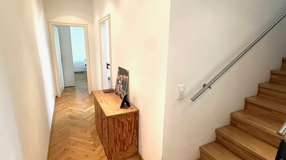 Schmaler, heller Flur in einem Wohnhaus mit weißen Wänden, eingelassenen Deckenleuchten und Fischgrät-Holzboden. Rechts führt eine Holztreppe mit Metallhandlauf nach oben, links stehen Türen zu angrenzenden Räumen und ein niedriger Holzschrank an der Wand.