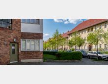 Straßenansicht eines Wohngebiets mit Reihenhäusern im traditionellen Baustil, gekennzeichnet durch Klinker- und Putzfassaden sowie rote Schrägdächer. Im Vordergrund sind gepflegte Grünflächen mit kleinen Bäumen sowie geparkte Autos zu sehen.