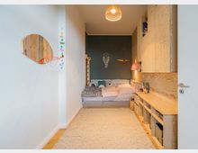 Kinderzimmer mit Bett und Holzschränken. Der Raum verfügt über eine grüne Akzentwand, eine Hängelampe aus Geflecht, eine kleine Basketballkorb-Dekoration und Spielzeugregale auf der rechten Seite.