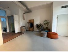 Minimalistisch eingerichteter Raum mit einer weißen Wand, einem Betonboden und einer kleinen Holzfläche. Zu den sichtbaren Möbeln gehören ein Kleiderständer, eine Holzfass-Dekoration, bunte Sitzpuffs, ein Pflanzengefäß sowie ein Faltparavent mit geometrischem Muster.