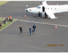 Ein kleines weißes Flugzeug steht auf einem asphaltierten Flugfeld, mit einer geöffneten Tür und zwei Personen in Geschäftskleidung an der Treppe. Im Vordergrund befindet sich ein Kamerateam, während links auf einer Wiese mehrere Personen und Ausrüstungen sichtbar sind.