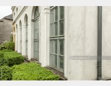 Seitenansicht eines historischen Gebäudes mit weißen Wänden, großen bogenförmigen Fenstern und angrenzendem gepflegten grünen Buschwerk. Im Hintergrund sind ein graues Nebengebäude und blühende weiße Rosen zu sehen.