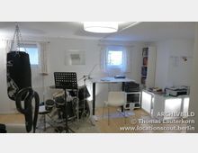 Ein multifunktionaler Raum mit Schlagzeug, Notenständer, Boxsack, Schreibtisch und Büchern in einem Regal. Der Raum hat weiße Wände, helle Vorhänge vor kleinen Fenstern und eine schlichte Beleuchtung.