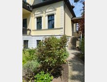 Hausfassade mit gelber Putzfassade und grünen Fensterrahmen im klassizistischen Stil, verziert mit Säulen und Rundbogenornamenten. Im Vordergrund ein Garten mit Sträuchern und einem gepflasterten Weg, der zu einer seitlichen Treppe führt.