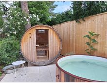 Holzbefeuerte Fasssauna mit Glastür in einem Garten, umgeben von blühenden Sträuchern und Bäumen, daneben ein kleines Rundbecken aus Holz mit Wasser sowie ein heller Holzzaun im Hintergrund.