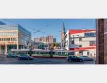 Straßenszene mit moderner Bebauung: Im Vordergrund fährt eine Straßenbahn entlang einer Kreuzung, umgeben von Autos. Im Hintergrund sind mehrstöckige Bürogebäude, ein modernes Gebäude mit roten Akzenten und eine Kirche mit spitzem Kirchturm zu sehen.