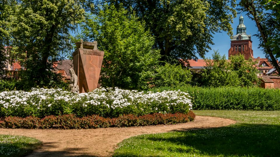 Gepflegter Stadtpark mit geschwungenen Sandwegen, großen Laubbäumen und einem runden Blumenbeet mit weißer Blüte um ein steinernes Denkmal. Im Hintergrund sind rote Dächer und ein Kirchturm mit grüner Kuppel zu sehen.