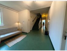 Ein schlichter Flur in einem Gebäude mit grünem Boden, weißen Wänden und einer Holzbank mit grauem Bezug auf der linken Seite. In der Mitte führt eine dunkle Treppe nach oben, und am Ende des Flurs ist eine Tür, durch die Tageslicht hereinfällt.