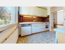 Helle Küche mit weiß-beigen Einbauküchenmöbeln, brauner gemusterter Fliesenrückwand und einem gefliesten Boden in Mosaikoptik. Links ein großes Fenster mit Blick auf einen Garten, rechts eine offene Verbindung zu einem angrenzenden Raum.