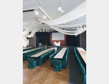 Ein Veranstaltungsraum mit einer Bühne im Hintergrund, langen Holztischen und Stühlen mit grünen Hussen. Der Raum ist mit weißen Stoffbahnen und Girlanden an der Decke dekoriert, und die Parkettbodenfläche ist sichtbar.