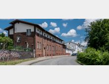 Eine gepflasterte Straße führt an einem Backsteingebäude mit weißen und braunen Akzenten entlang, das von einem Zaun und etwas Grün umgeben ist; im Hintergrund sind weiße Gebäude sichtbar. Rechts der Straße befindet sich eine bewachsene Grünfläche, während der Himmel klar und von vereinzelten Wolken bedeckt ist.