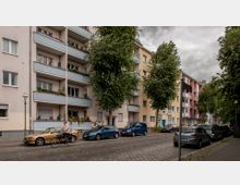 Mehrstöckige Wohngebäude mit Balkonen in Pastellfarben entlang einer ruhigen gepflasterten Straße, gesäumt von parkenden Autos und großen Bäumen; eine Frau fährt Fahrrad auf der Straße.